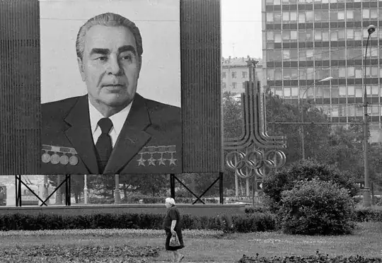 A black and white photo showing a large billboard of Leonid Brezhnev, with what looks to be a large steel sculpture of the 1980 Summer Olympics emblem in the background.