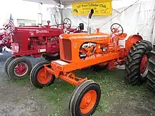 Two tractors on display in a tent.  In the rear, a red one labeled "McCORMICK FARMALL".  In front is an orange one, labeled ALLIS-CHALMERS with a "WD 45" emblem.  A banner behind them reads CNY Two-Cylinder Club"