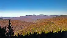 A landscape of forested mountains, in autumn color, underneath a blue sky, dominated by three rounded peaks in the distance with dark tops