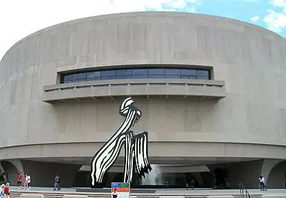 A large, convexly curving, 3 story tall, gray wall punctured on the second floor by a long strip of windows and a short balcony. A large black and white sculpture of a paint stroke sits in front of the building, stretching from the ground to the balcony