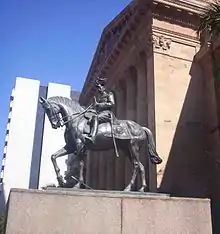 Equestrian statue in dark grey metal of George&nbsp;V in military dress uniform on a plinth of red granite outside a Classical building of red sandstone