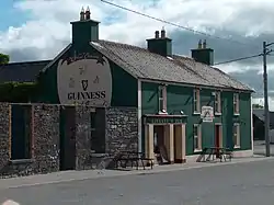 Photo of Linnane's Pub - A green colour building with "Linnane's Pub" written in light yellow above the main door. The door and window frames are light yellow. There is a large advertisement for Guinness on the side of the building. There are two park benches in front of the pub. There is a grey structure on the left side of the pub. The photo was taken during the day when the sky was blue with white clouds. It is taken from outside, and from opposite side of the road