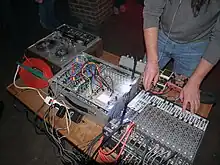 A photo of Pete Swanson (only lower part of his body is visible) performing live, with various pieces of electronic equipment placed on a table in front of him.