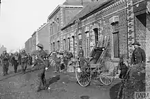 Crosses painted on the building denote this as an aid station. Soldiers stand around outside the building, while others are matching past.