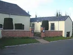 Two single-storey cream buildings with slate roofs at an angle to each other. On the left is part of the chapel; on the right is the assembly hall with two sash windows.  In front is a brick wall and gateposts; above the gate is an overthrow with a lantern.