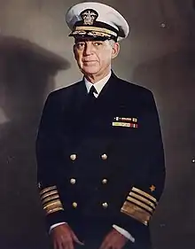 Man in dark blue suit and tie, wearing peaked cap and two rows of ribbons